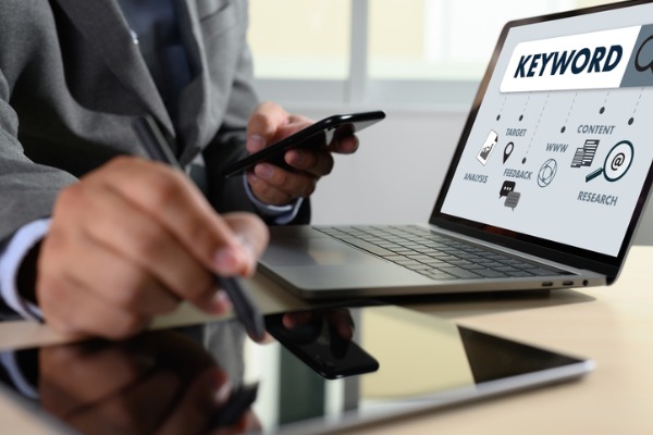 An image showing someone working on a smartphone and tablet while his laptop shows a keyword search. 